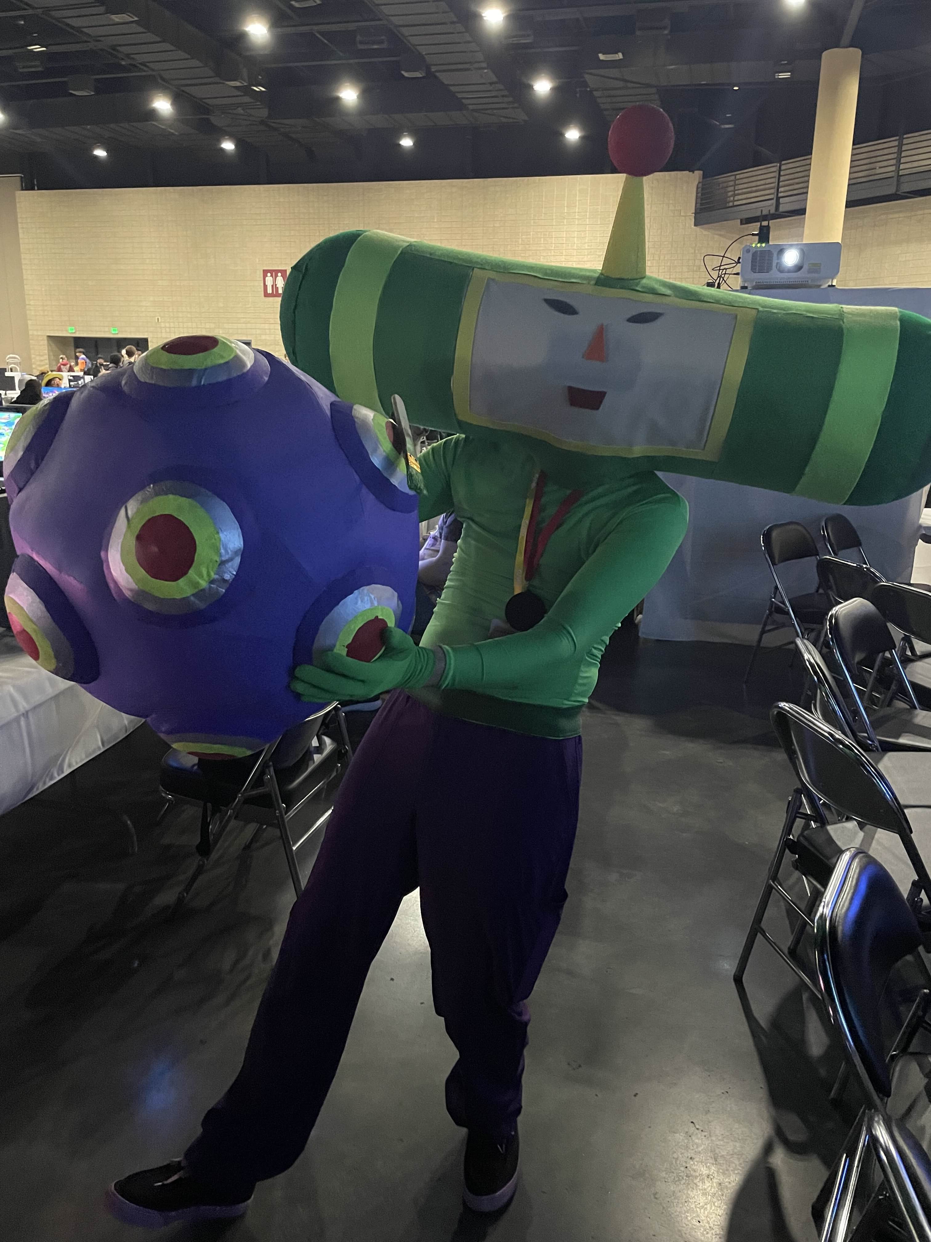 a guy dressed up in a cosplay of the prince from katamari damacy. he is holding a katamari with some things stuck to it.